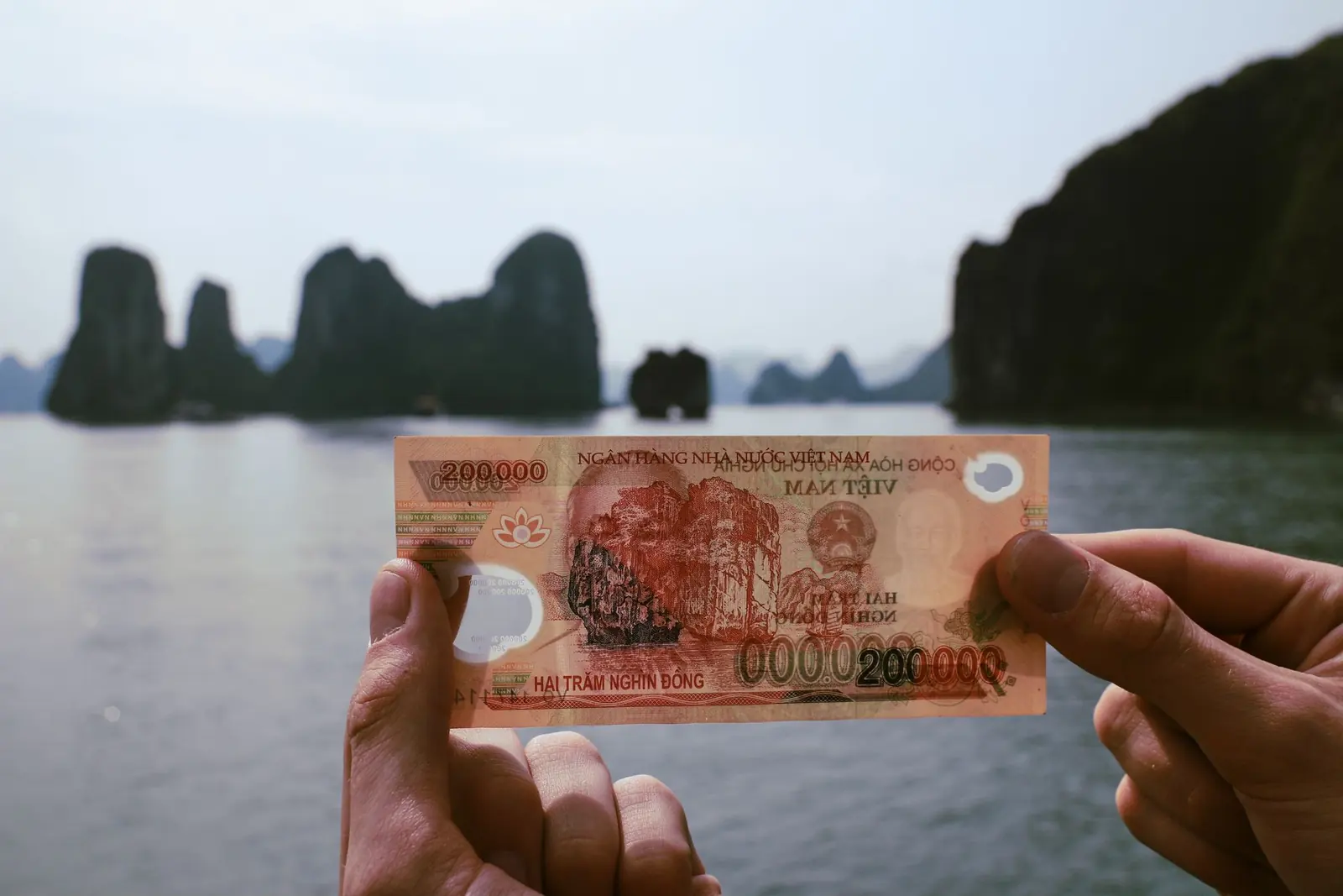 Bàn tay cầm tờ tiền hai trăm nghìn đồng trước vịnh Hạ Long